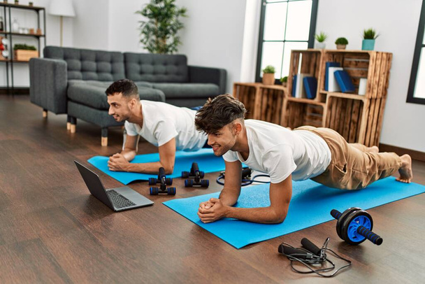 Dois homens hispânicos casal sorrindo confiante ter abdominais on-line aula de exercício em casa - Foto, Imagem