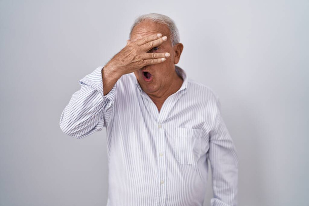 Senior man with grey hair standing over isolated background peeking in shock covering face and eyes with hand, looking through fingers with embarrassed expression.  - Fotografie, Obrázek