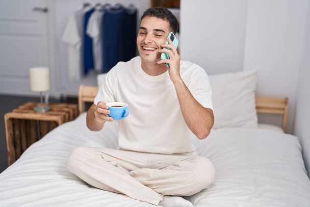Junger hispanischer Mann telefoniert mit Smartphone und trinkt Kaffee im Schlafzimmer - Foto, Bild