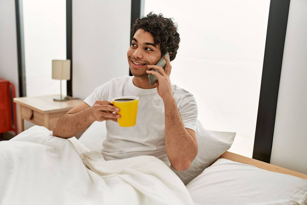 Hombre hispano joven bebiendo café y hablando en el teléfono inteligente en el dormitorio. - Foto, Imagen