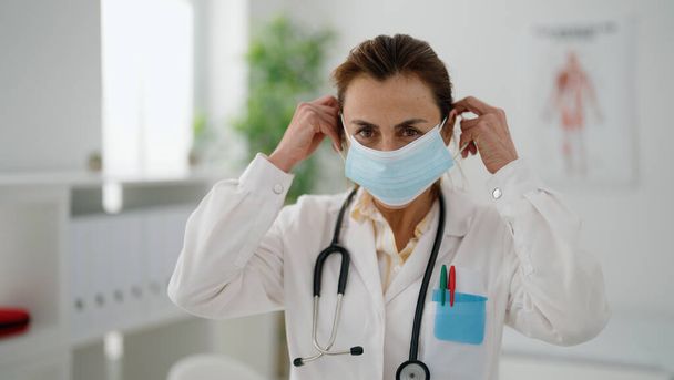 Middle age hispanic woman wearing doctor uniform and medical mask at clinic - Foto, imagen
