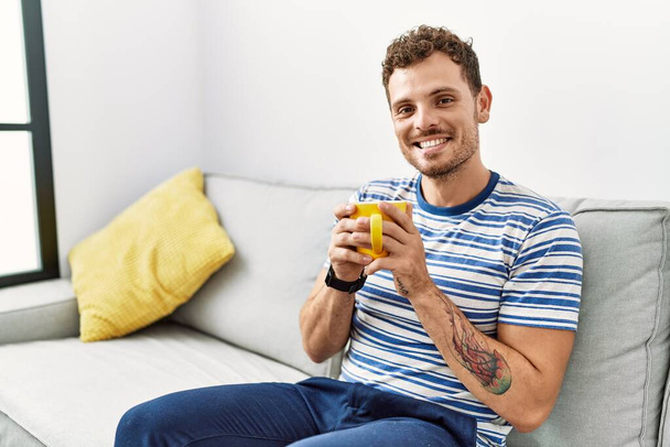 Молодой латиноамериканец, уверенно улыбающийся, пьющий кофе дома - Фото, изображение