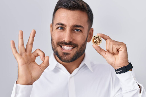 Przystojny Hiszpan trzyma kardano kryptowaluta monety robi ok znak palcami, uśmiechając przyjazny gest doskonały symbol  - Zdjęcie, obraz