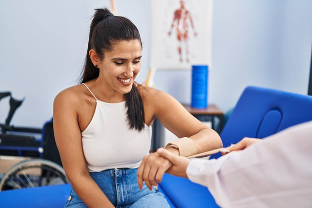 Young hispanic woman smiling confident bandaging wrist at physiotherapy clinic - Photo, Image