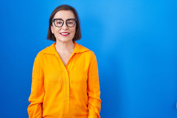 Латиноамериканка средних лет в очках, стоящих на синем фоне со счастливой и прохладной улыбкой на лице. счастливчик.  - Фото, изображение
