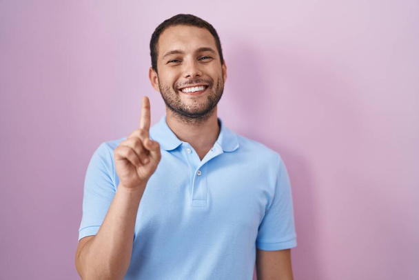 Hispánec stojící nad růžovým pozadím ukazující a ukazující prstem číslo jedna a usmívající se sebevědomě a šťastně.  - Fotografie, Obrázek