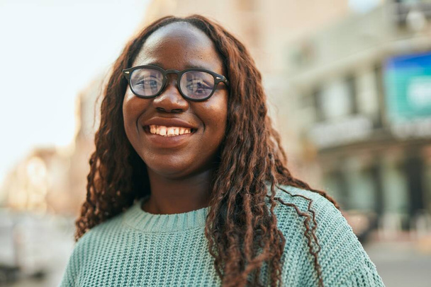 Νεαρή Αφρικανή γυναίκα χαμογελά ευτυχισμένη φορώντας γυαλιά στην πόλη - Φωτογραφία, εικόνα