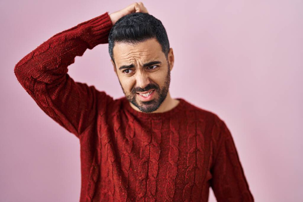 Young hispanic man with beard wearing casual sweater over pink background confuse and wonder about question. uncertain with doubt, thinking with hand on head. pensive concept.  - Photo, Image