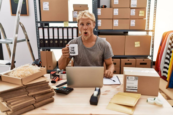 Jonge blonde man drinken koffie werken bij online winkel bang en verbaasd met open mond voor verrassing, ongeloof gezicht  - Foto, afbeelding