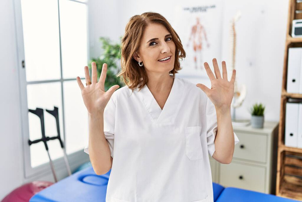 Fisioterapeuta mujer de mediana edad que trabaja en la clínica de recuperación del dolor mostrando y señalando con los dedos número diez mientras sonríe confiado y feliz.  - Foto, imagen