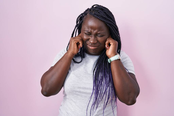 Молодая африканская женщина, стоящая на розовом фоне, закрывает уши пальцами с раздраженным выражением для шума громкой музыки. Концепция deaf.  - Фото, изображение