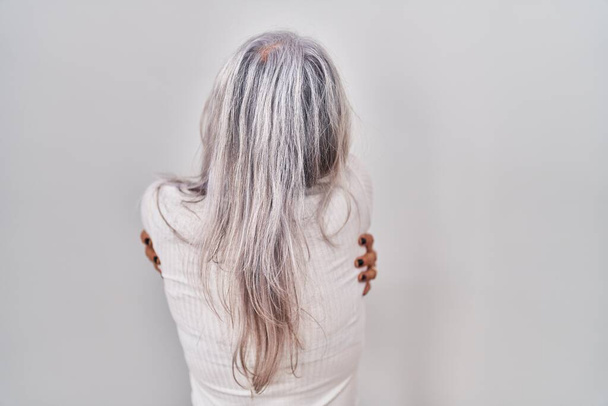 Žena středního věku s šedivými vlasy stojící nad bílým pozadím objímající se šťastně a pozitivně pozpátku. self love a self care  - Fotografie, Obrázek