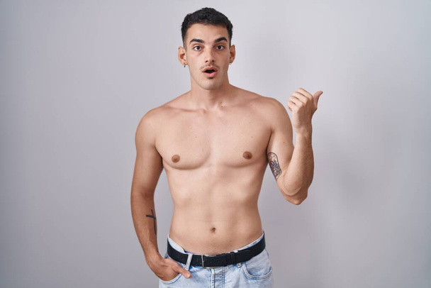 Knappe Latijns-Amerikaanse man staat shirtloos verrast wijzend met de hand vinger naar de zijkant, open mond verbaasd uitdrukking.  - Foto, afbeelding