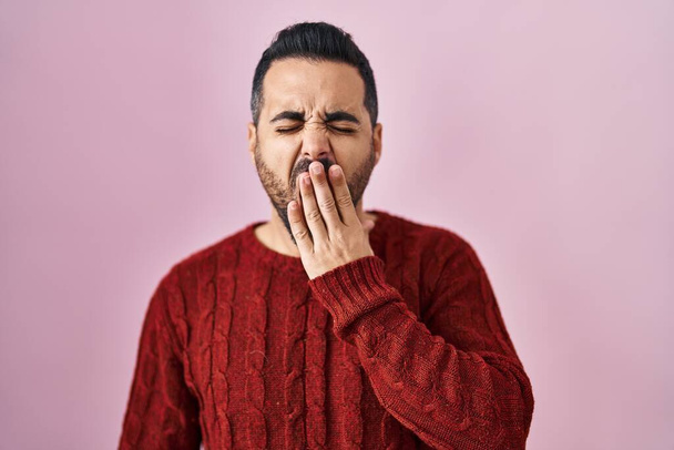 Joven hispano con barba vistiendo suéter casual sobre fondo rosa aburrido bostezando cansado cubriendo la boca con la mano. inquietud y somnolencia.  - Foto, imagen