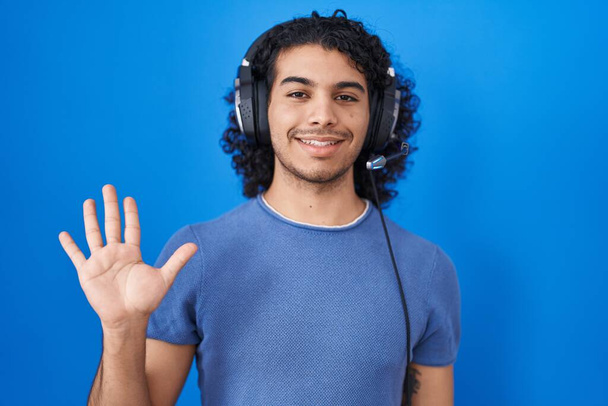 Latinomies, jolla on kiharat hiukset kuunnellen musiikkia kuulokkeilla, jotka näyttävät ja osoittavat sormilla numero viisi hymyillen itsevarmoina ja onnellisina.  - Valokuva, kuva