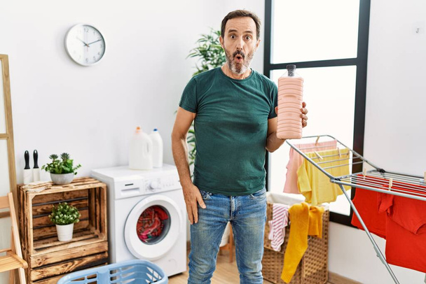 Muž středního věku s plnovousem praní prádla držení saponátu láhev vyděšený a ohromený s otevřenými ústy pro překvapení, nevěřící tvář  - Fotografie, Obrázek