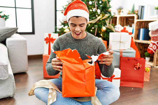 Jeune homme hispanique déballer cadeau assis par arbre de Noël à la maison - Photo, image