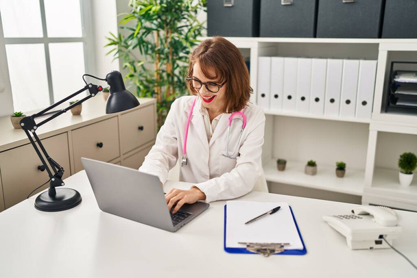 Young caucasian woman wearing doctor uniform using laptop at clinic - Фото, изображение
