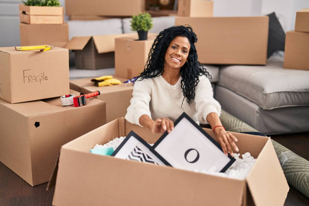 Африканская американка распаковывает картонную коробку, сидя на полу в новом доме - Фото, изображение