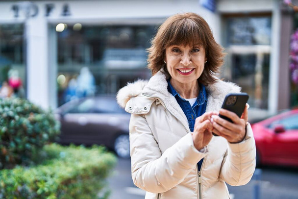 Sokakta akıllı telefon kullanan orta yaşlı bir kadın gülümsüyor. - Fotoğraf, Görsel