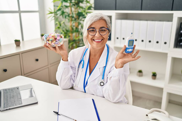 Kobieta w średnim wieku z siwymi włosami nosząca mundur lekarza trzymająca monitor glukozy uśmiechnięta ze szczęśliwym i chłodnym uśmiechem na twarzy. wykazujące zęby.  - Zdjęcie, obraz