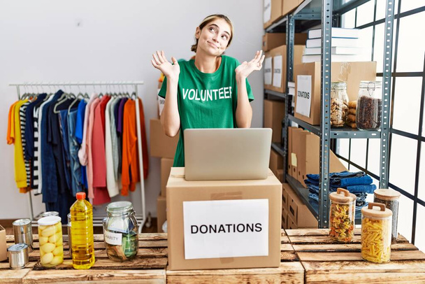 Νεαρή ξανθιά γυναίκα που φοράει εθελοντική μπλούζα σε δωρεές στέκεται ανίδεη και συγκεχυμένη έκφραση με χέρια και χέρια σηκωμένα. έννοια της αμφιβολίας.  - Φωτογραφία, εικόνα