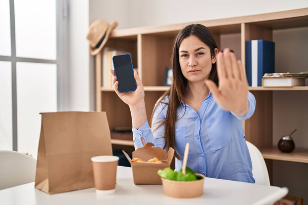 Young brunette woman eating take away food at home showing smartphone screen with open hand doing stop sign with serious and confident expression, defense gesture  - 写真・画像