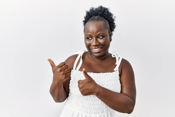 Mladá africká žena stojící nad bílým izolovaným pozadím ukazuje na zadní stranu s rukou a palci nahoru, s úsměvem sebevědomě  - Fotografie, Obrázek