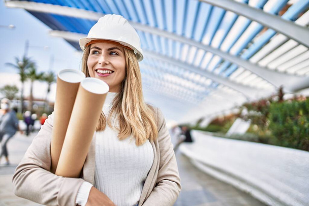 Young blonde woman wearing hardhat holding blueprints at street - 写真・画像
