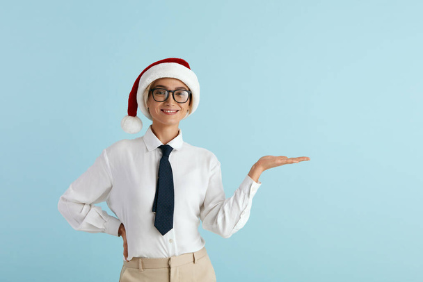 Smiling Businesswoman Pointing Hand. Positive Caucasian Lady in Santa Hat Pointing Away Paying Your Attention at Empty Space for Advertisement. Indoor Studio Shot Isolated on Blue Background  - Foto, imagen