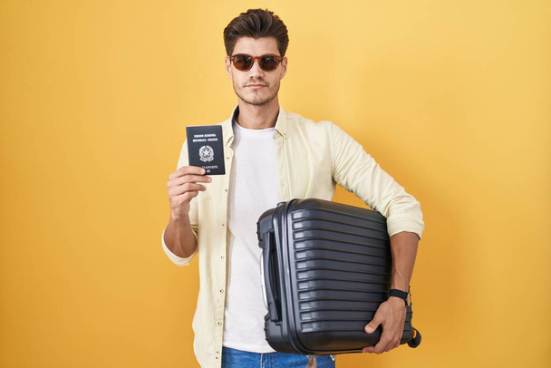 Joven hombre hispano sosteniendo maleta que va de vacaciones de verano con pasaporte italiano relajado con expresión seria en la cara. simple y natural mirando a la cámara.  - Foto, Imagen