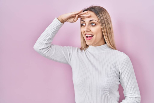 Young blonde woman wearing white sweater over pink background very happy and smiling looking far away with hand over head. searching concept.  - Fotografie, Obrázek