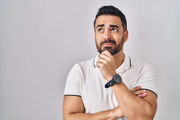 Jeune homme hispanique avec barbe portant des vêtements décontractés sur fond blanc pensant inquiet d'une question, préoccupé et nerveux avec la main sur le menton  - Photo, image