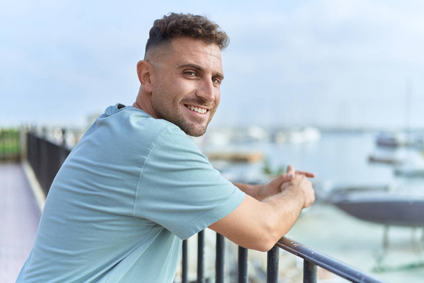 Jeune homme hispanique souriant confiant appuyé sur la balustrade au bord de la mer - Photo, image