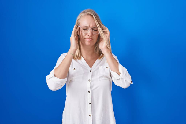 Young caucasian woman standing over blue background with hand on head for pain in head because stress. suffering migraine.  - 写真・画像