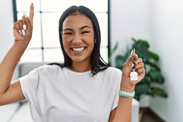 Young hispanic woman holding keys of new home smiling amazed and surprised and pointing up with fingers and raised arms.  - Photo, Image