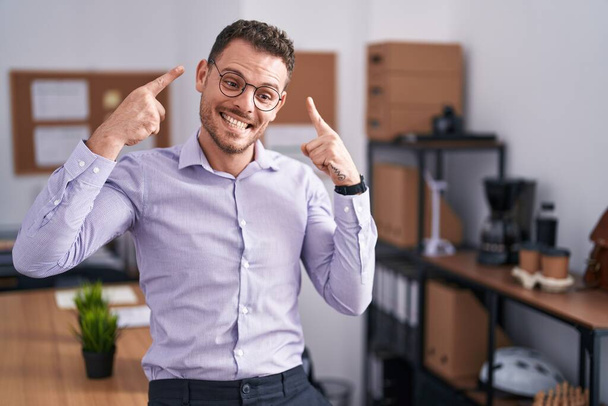 Jeune homme hispanique au bureau souriant pointant vers la tête avec les deux doigts de la main, bonne idée ou pensée, bonne mémoire  - Photo, image