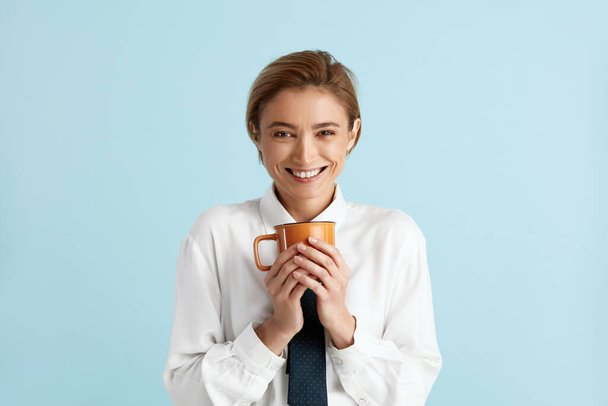 Smiling Businesswoman Holding Coffee. Portrait of Friendly Young Adult Woman Drinking Coffee, Holding Drink in Cup and Smiling. Indoor Studio Shot Isolated on Blue Background  - Photo, image