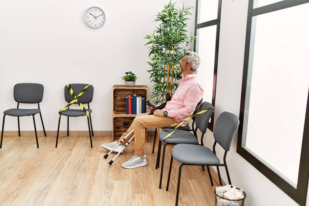 Middle age grey-haired man injured sitting on chair holding crutches at clinic waiting room - Photo, Image
