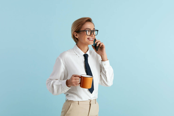 Деловая женщина, держащая кофе. Портрет дружелюбной молодой женщины в очках, говорящей по мобильному телефону, держащей напиток в кубке и улыбающейся. Крытая студия, снятая на голубом фоне  - Фото, изображение