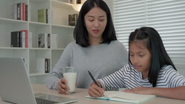 Asian young little girl learn at home. Do homework with kind mother help, encourage for exam. Mom pass on a glass of milk to daughter. Girl happy Homeschool. Mom teach and advice education together. - Footage, Video