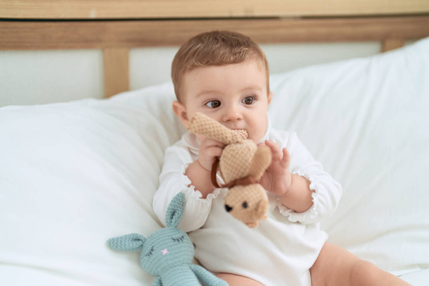 Adorable toddler sitting on bed bitting doll at bedroom - Foto, afbeelding