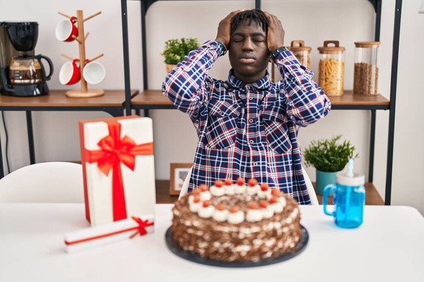 African man with dreadlocks celebrating birthday holding big chocolate cake suffering from headache desperate and stressed because pain and migraine. hands on head.  - Photo, image