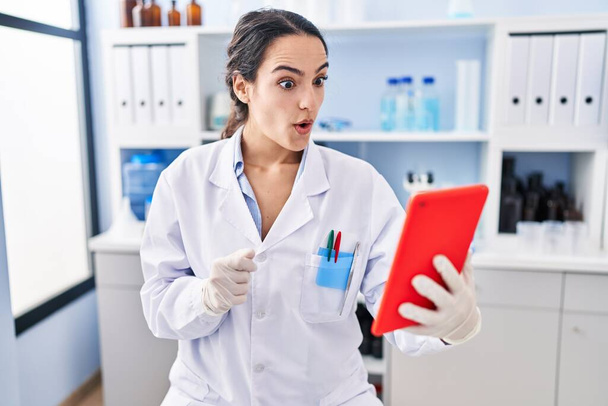 Young brunette woman working at scientist laboratory doing online call scared and amazed with open mouth for surprise, disbelief face  - Zdjęcie, obraz