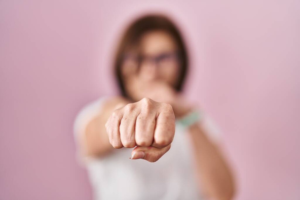 Middle age hispanic woman standing over pink background punching fist to fight, aggressive and angry attack, threat and violence  - Фото, изображение