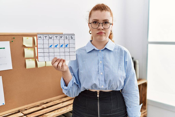Junge rothaarige Frau mit Reisekalender im Büro denkt Haltung und nüchternen Ausdruck sieht selbstbewusst aus  - Foto, Bild