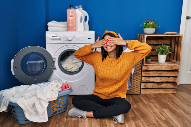 Young hispanic woman doing laundry covering eyes with hands smiling cheerful and funny. blind concept.  - Valokuva, kuva