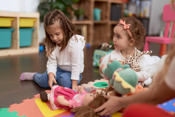Две маленькие девочки играют в куклы, сидя на полу в детском саду - Фото, изображение