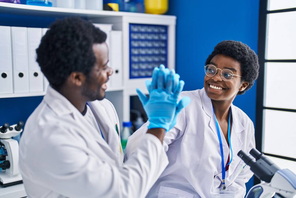 African american man and woman scientists high five with hands raised up at laboratory - Foto, afbeelding