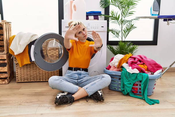 Çamaşır makinesinin yanında çamaşır yıkayan, elleri ve parmakları kullanarak çerçeve yapan genç sarışın kadın, kamera açısı.  - Fotoğraf, Görsel
